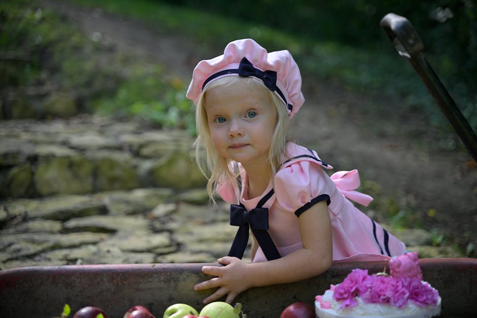 A young girl in a pink dress and bonnet poses with apples and flowers outdoors.