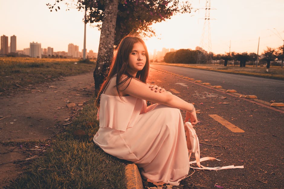 A young woman in a flowing dress sits by a city road at sunset, capturing a serene moment.