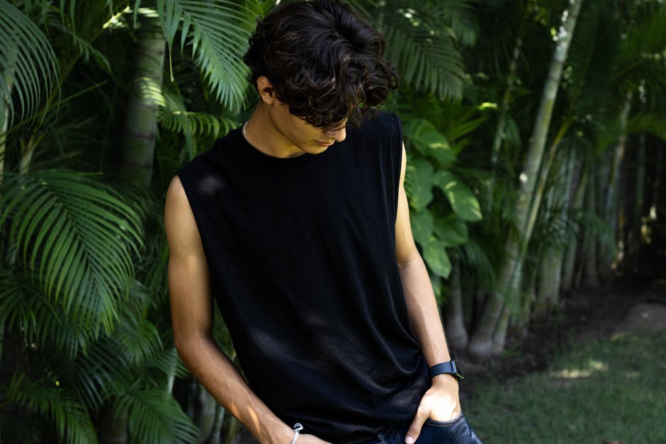 A young man in a sleeveless black shirt stands surrounded by lush green tropical plants.