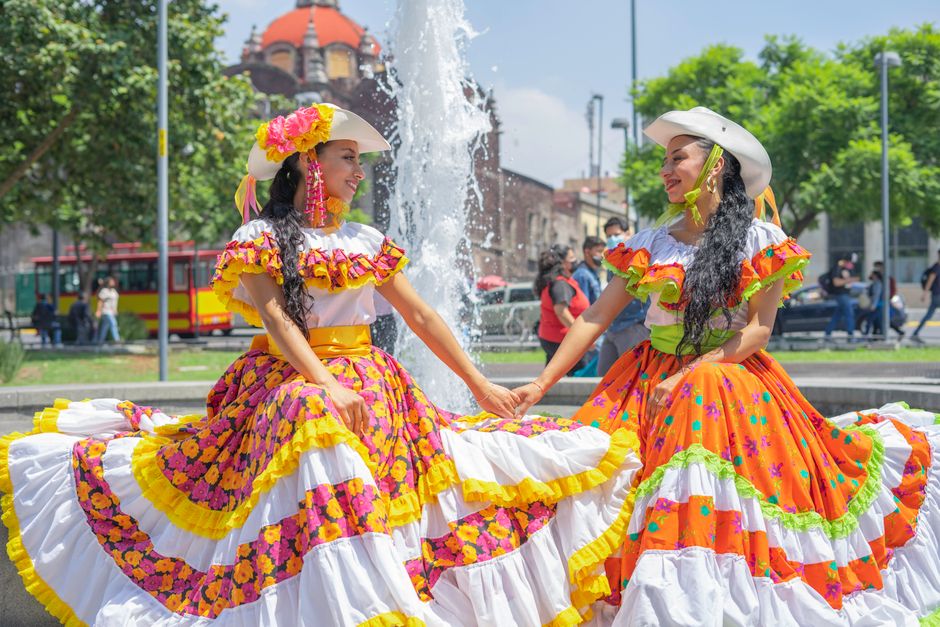 Two women in vibrant traditional Mexican dresses holding hands in front of a fountain.