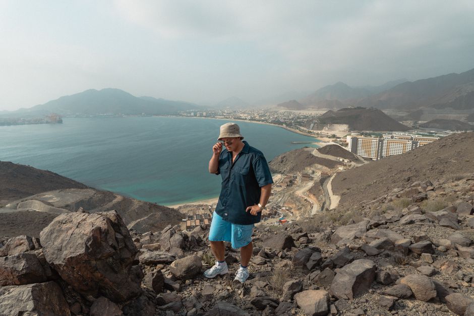 A person hiking on rocky terrain with panoramic views of Khor Fakkan's coastline and cityscape.
