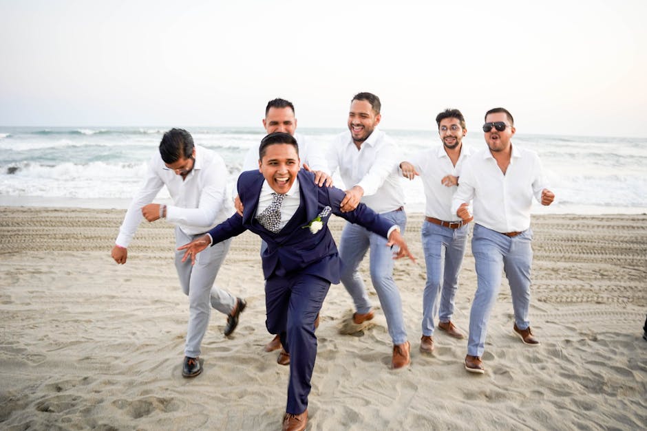 A joyful moment of a groom and his groomsmen running on a sandy beach during a wedding.